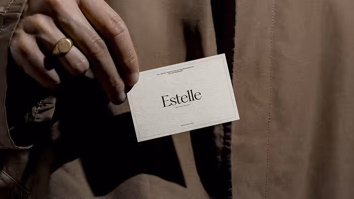 Hand holding a white business card with the name 'Estelle' printed in black serif font against a brown fabric background.